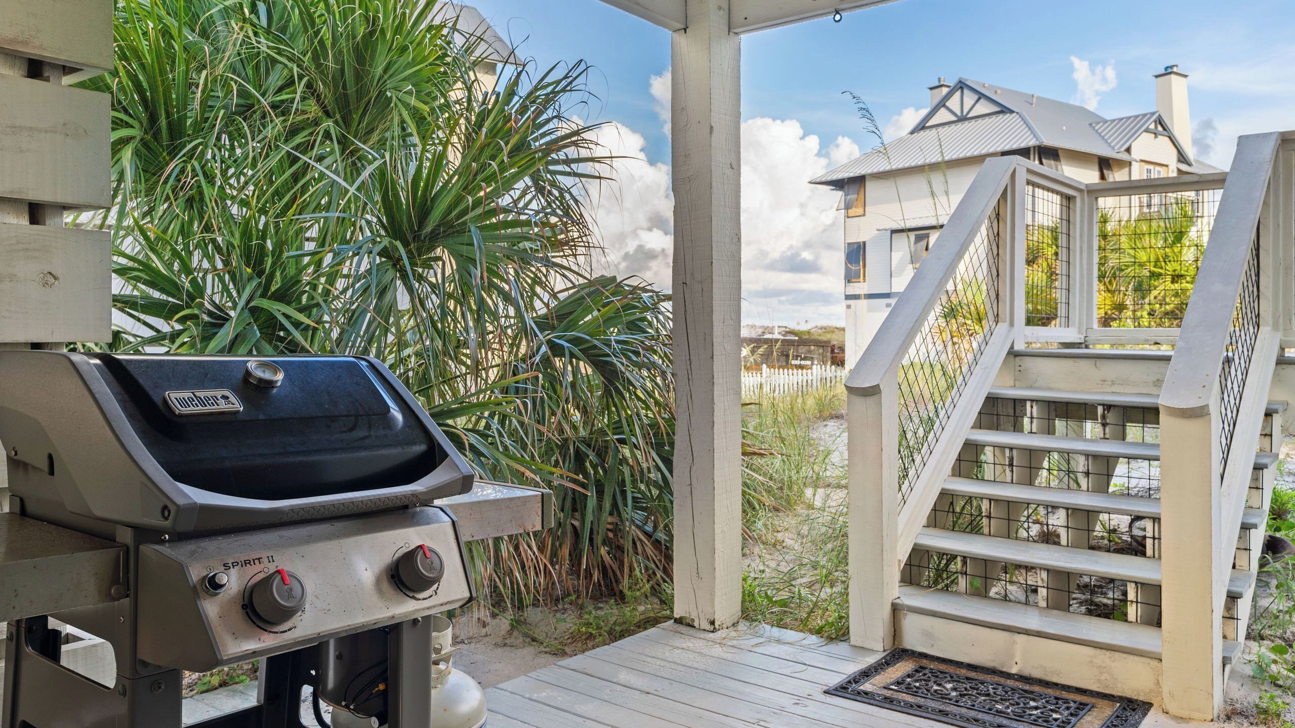 A barbecue grill, wooden deck, and a house with a white fence and stairs.