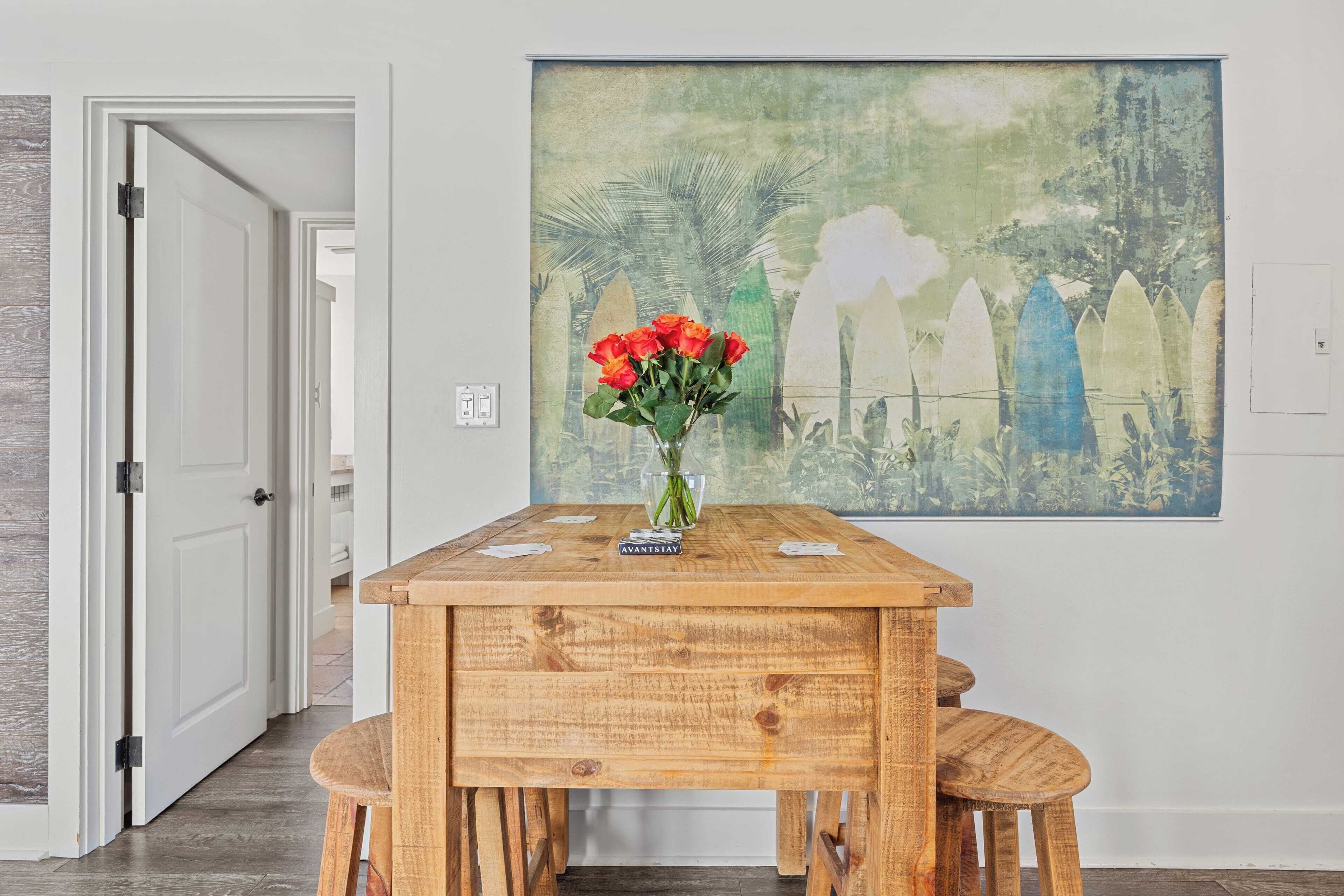 A wooden dining table with a vase of red flowers and a placemat, set against a wall with a large abstract painting.