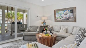 A modern living room with a large sliding glass door to a deck, a round wooden coffee table with snacks, a grey sofa, and a painting on the wall. - Stoked by Avantstay Gulf View, Steps to the Beach (Santa Rosa Beach)