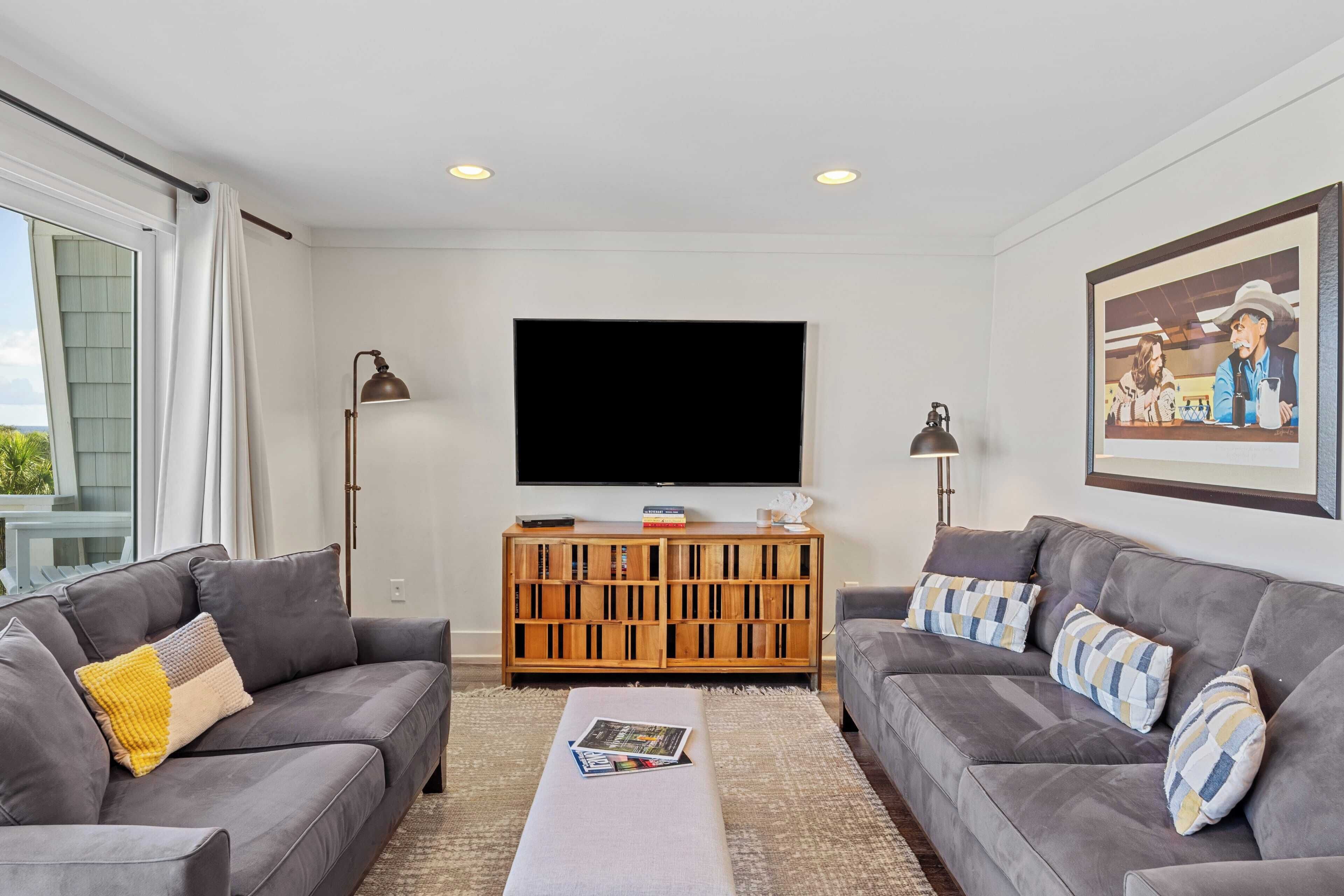 A modern living room with two grey sofas, a coffee table, a flat-screen TV, wooden entertainment unit, and a large window.