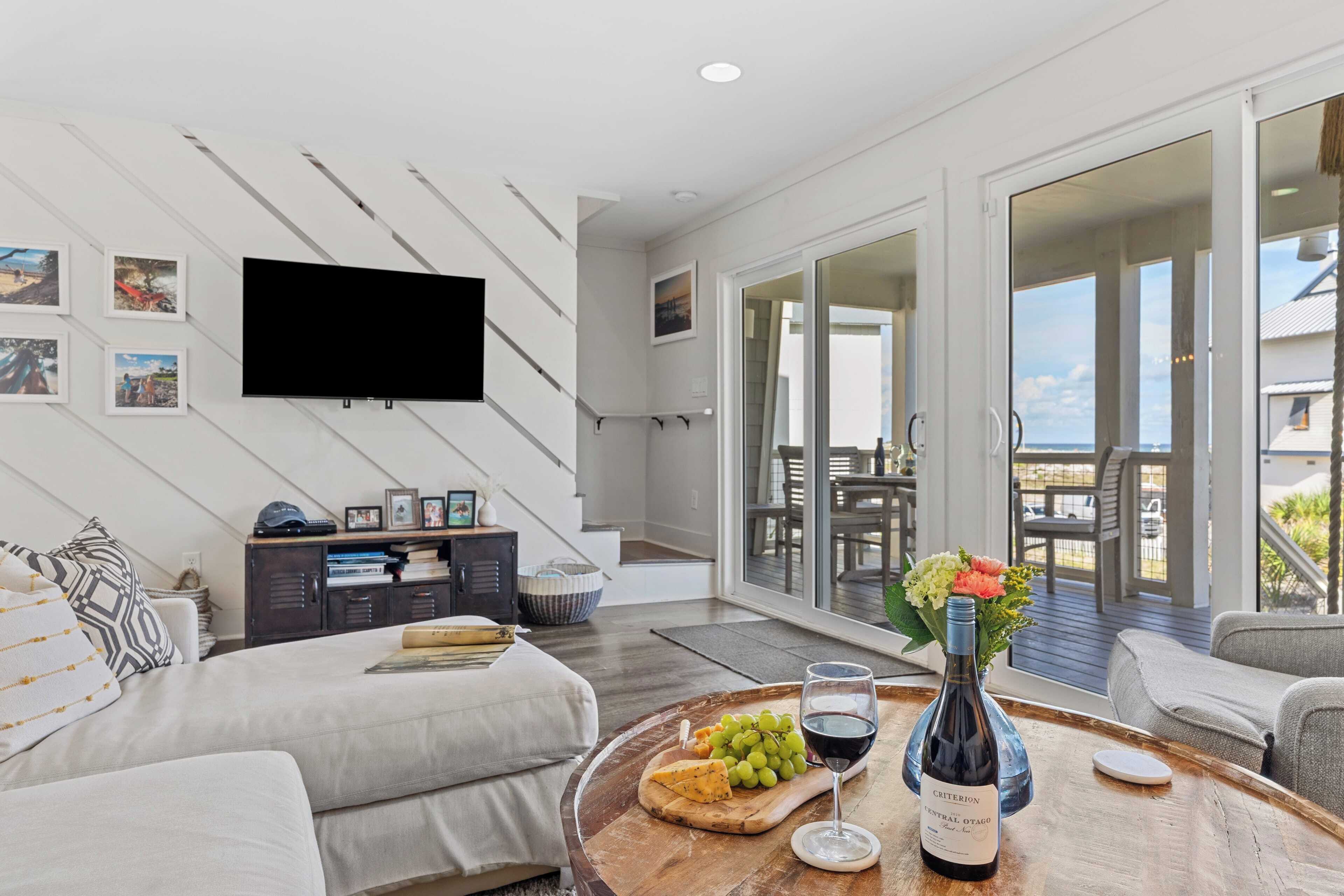 A modern living room with a large flat-screen TV, a wooden coffee table with a bottle of wine, grapes, and cheese, and a sofa.