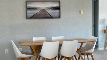 Zona pranzo con tavolo in legno e sedie bianche, un quadro appeso al muro e pavimento con tappeto.