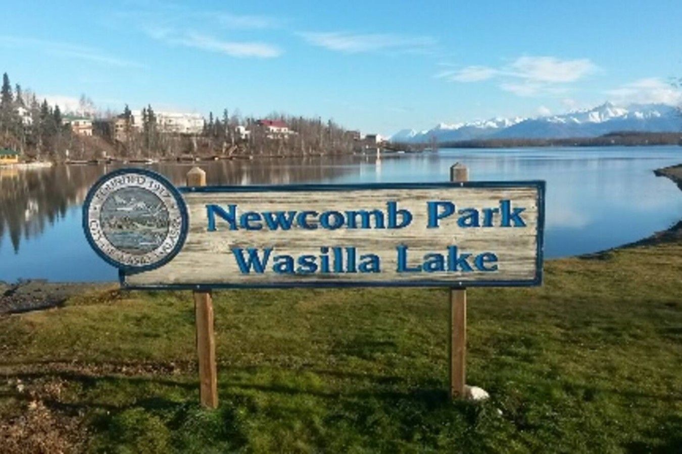 A wooden sign with 'Newcomb Park Wasilla Lake' written on it, set against a backdrop of a lake and mountains.