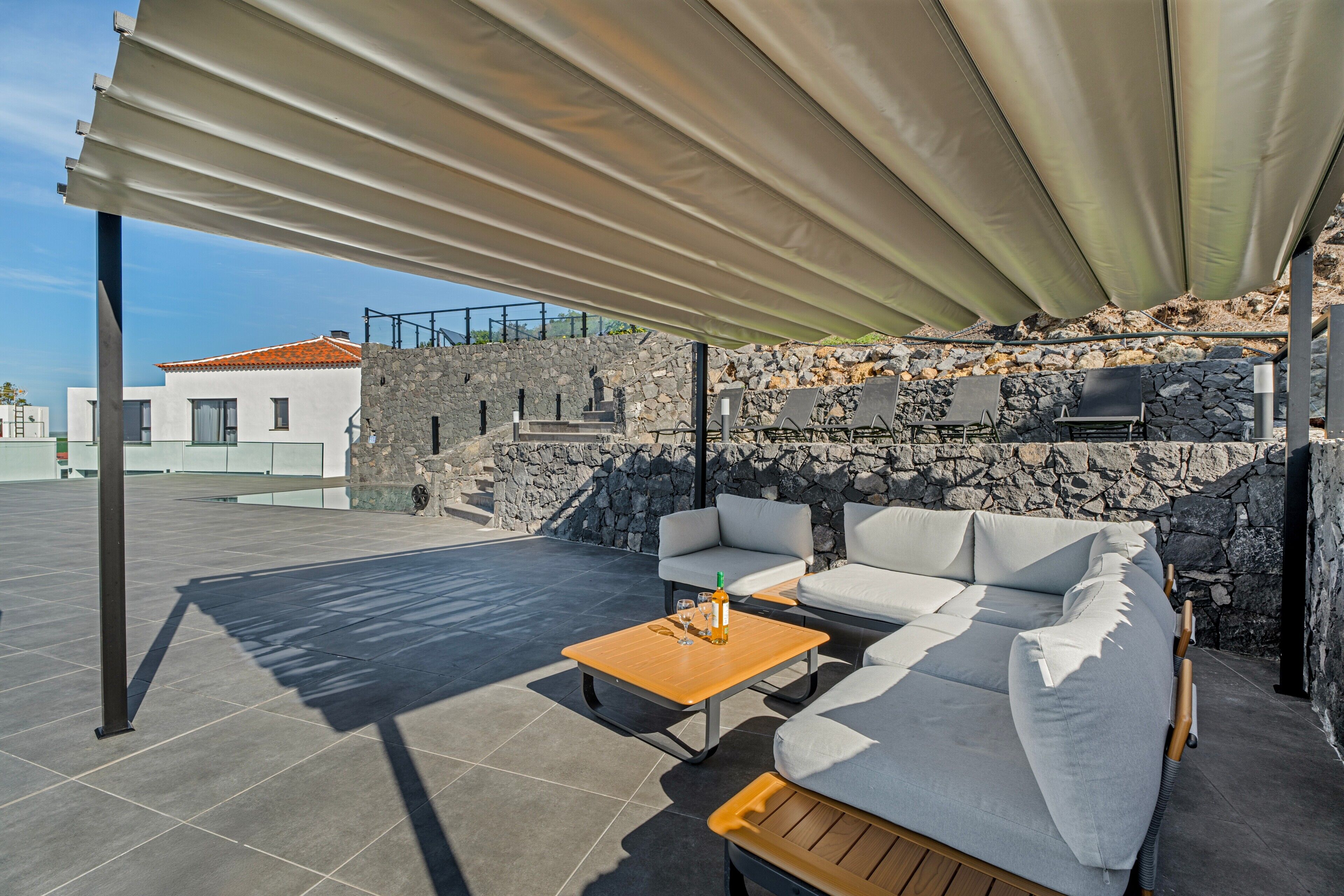 A covered outdoor seating area with a stone wall, a white building in the background, and a clear sky.