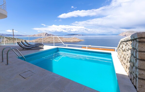 Una piscina con vistas al mar y a las montañas, barandilla blanca y muro de piedra.