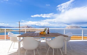 Una mesa de madera con sillas blancas en un balcón con vista a una masa de agua y formas de terreno distantes.