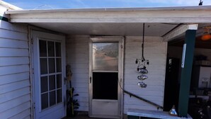 A small, single-story building with a white exterior and a green door. There is a small porch area with a metal railing and a hanging light fixture.