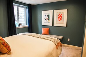 Une chambre à coucher avec un lit, une fenêtre avec des rideaux, une table de chevet et deux œuvres d’art encadrées accrochées au mur.