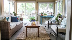 Ein gemütliches Wohnzimmer mit einem Rattan-Sofa, einem Holztisch und einem Sessel. Durch große Fenster ist der Garten zu sehen.