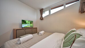 A bedroom with a TV on a wooden cabinet, two beds with towels, a window with curtains, and a view of a forest stream. - Modern home with pool and view (Popovaca)