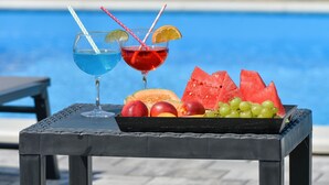Two drinks with straws and fruit garnishes, a watermelon slice, and a tray of fruits on a table by a pool. - Modern home with pool and view (Popovaca)