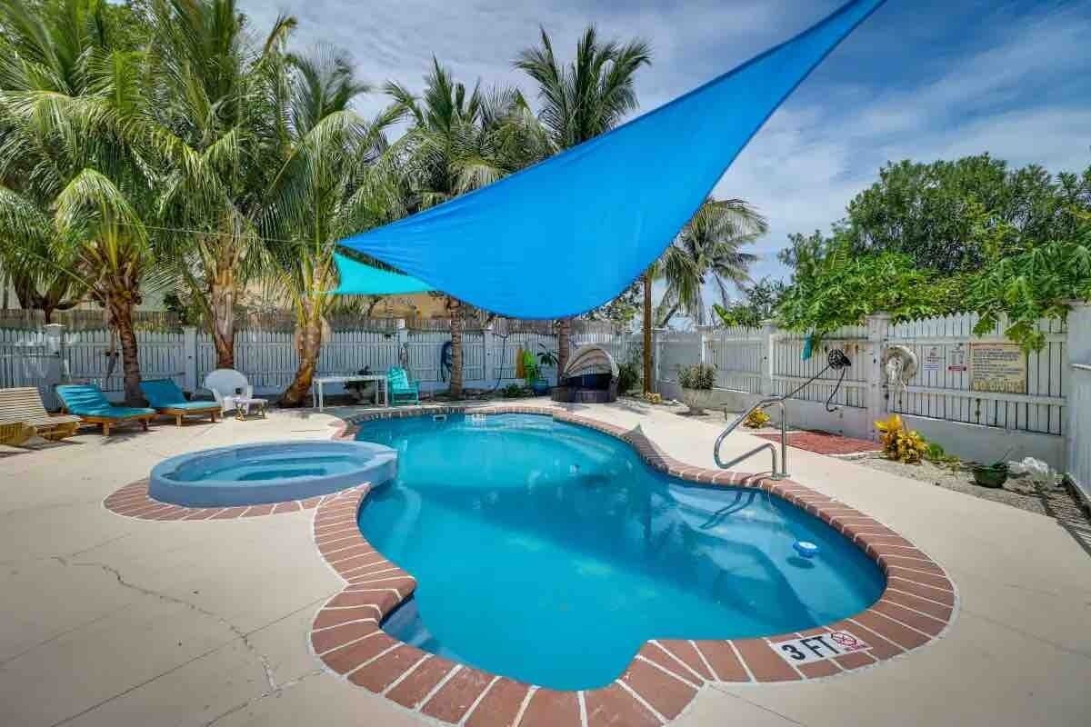 Une piscine bordée de briques, agrémentée de palmiers et surmontée d’une voile d’ombrage bleue.