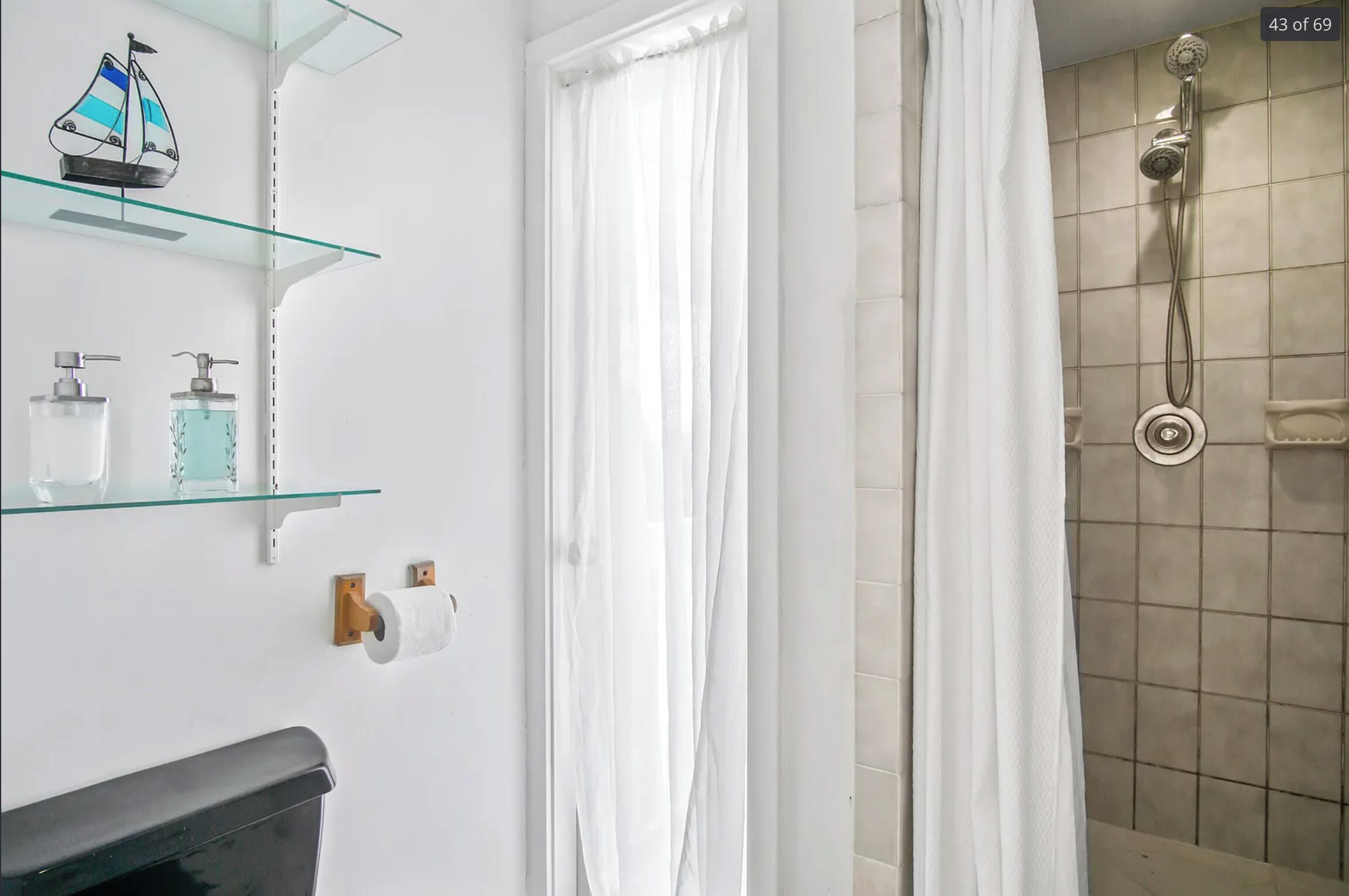 A bathroom with a glass shelf holding toiletries, a toilet paper holder, and a shower with a curtain.