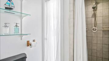 A bathroom with a glass shelf holding toiletries, a toilet paper holder, and a shower with a curtain.
