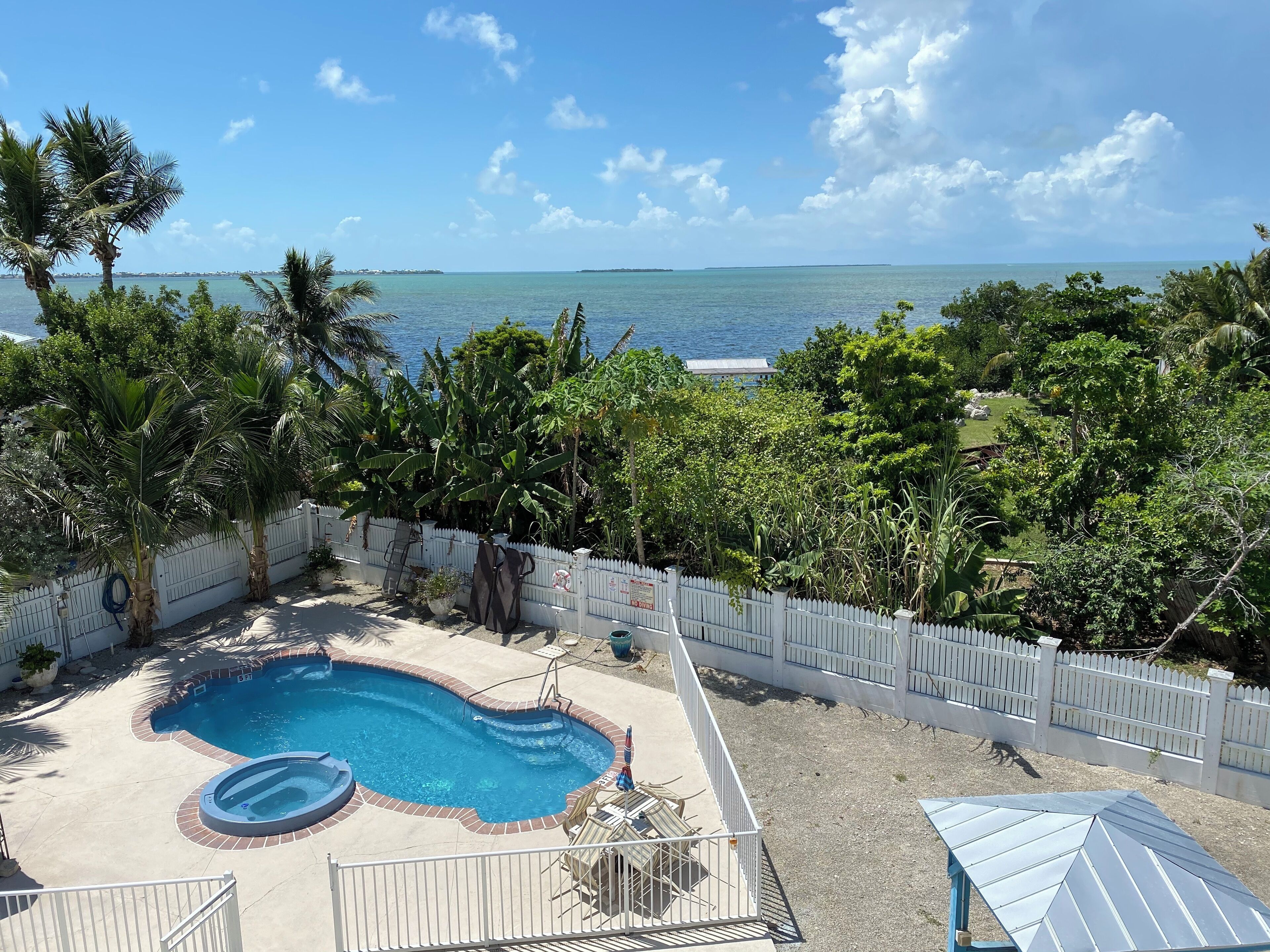 Un jardin avec une piscine, entouré d’une clôture blanche et d’une végétation luxuriante, offrant une vue imprenable sur une mer bleue et limpide.