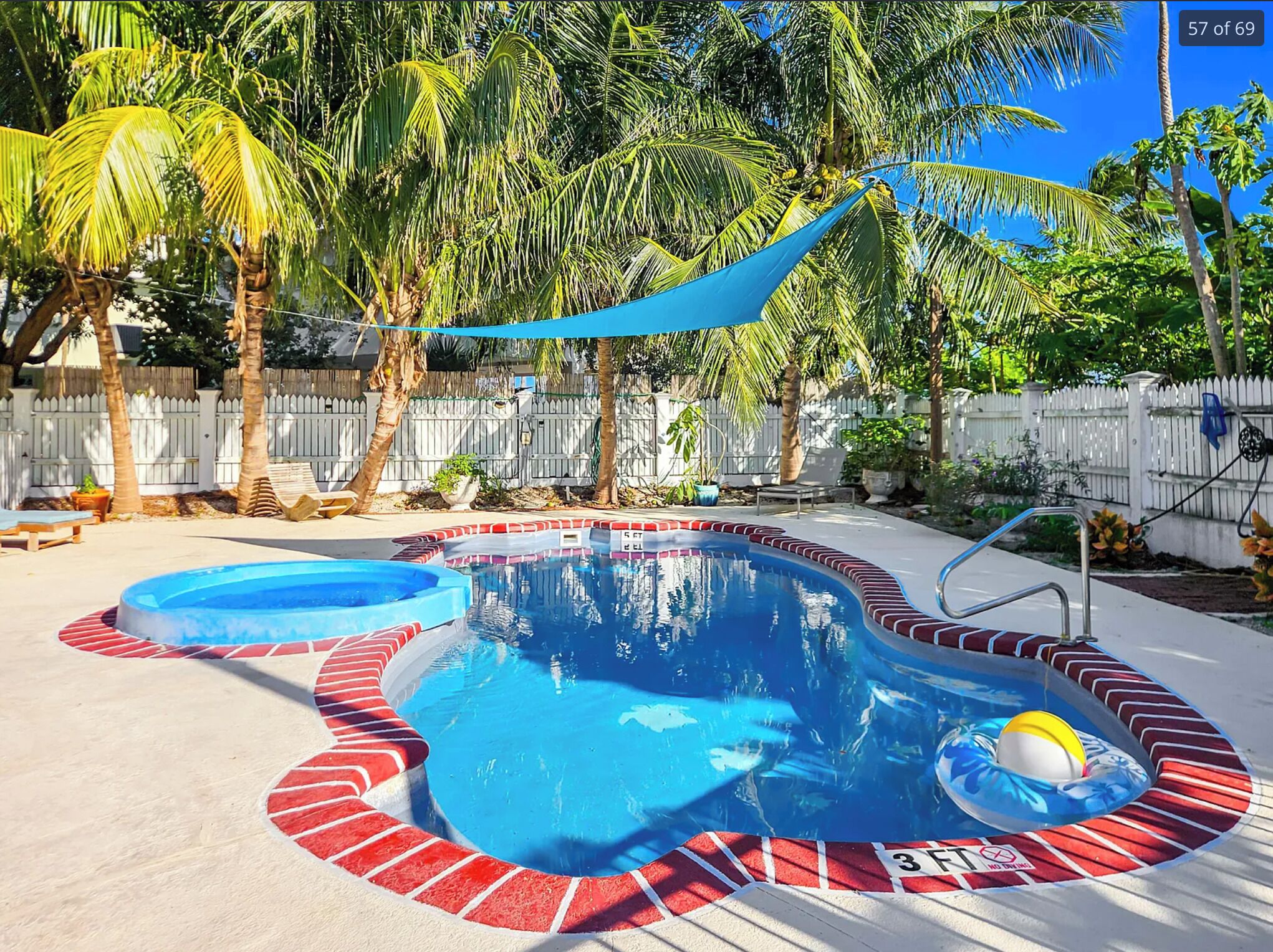 Ein Swimmingpool mit roter und weißer Umrandung, umgeben von Palmen und einem weißen Zaun.