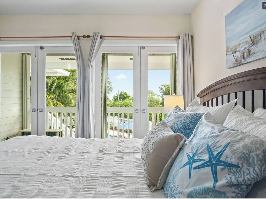 Ein Schlafzimmer mit einem großen Bett, weißer Bettwäsche und Blick auf einen Pool und Palmen durch offene Französische Türen.