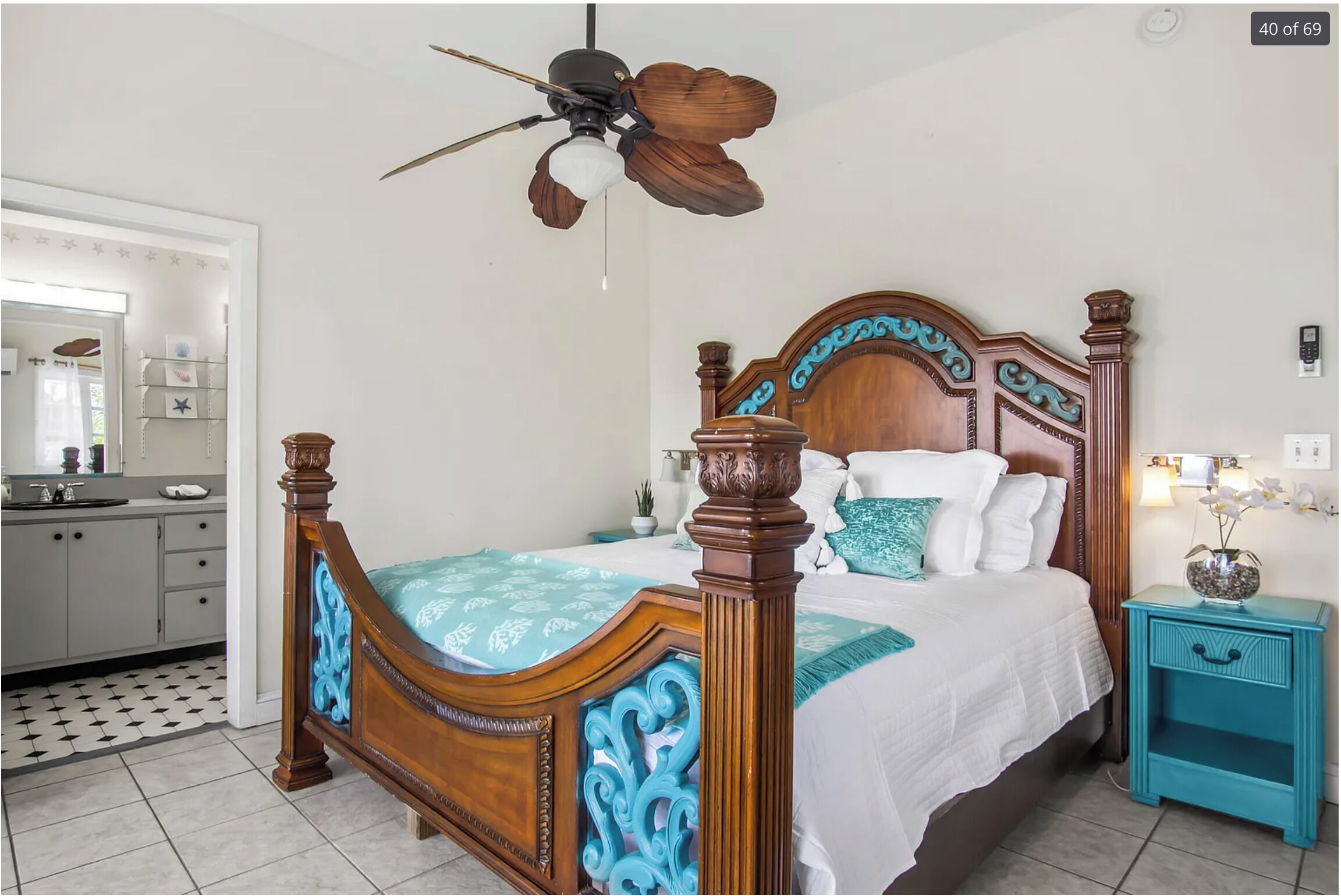 A bedroom with a wooden four-poster bed, a ceiling fan, a nightstand with a vase, and a bathroom visible in the background.
