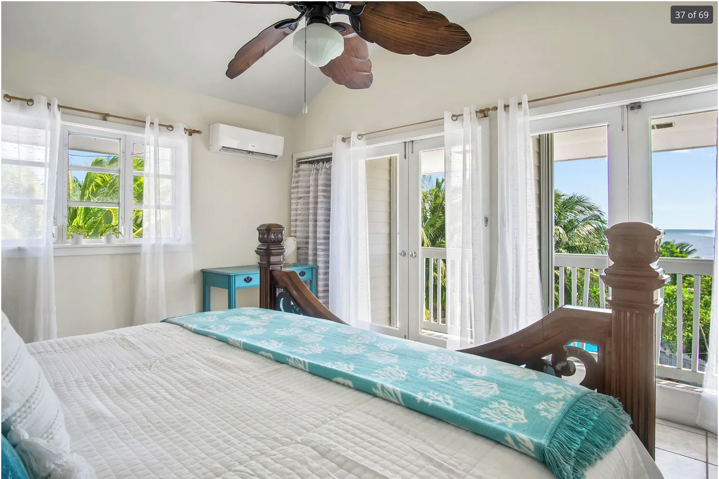Quarto com cama, ventoinha no teto, janela com cortinas, uma mesinha e vista para o exterior.