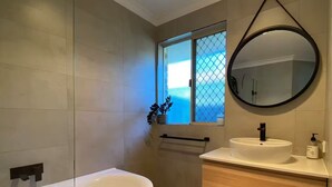 A modern bathroom with a freestanding bathtub, a round mirror, a sink with a wooden vanity, and a window with a view of the outside. - Cosy Home near SValley & Airport (Ashfield)