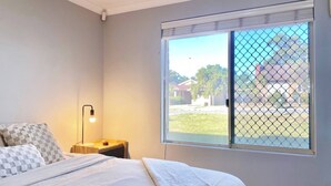 A bedroom with a bed, a nightstand, a lamp, and a window with a view of a residential area. - Cosy Home near SValley & Airport (Ashfield)