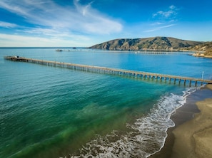 A coastal scene with a pier extending into calm waters, a sandy beach, and a hilly landscape in the background.
