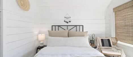 A bedroom with a bed, bedside table, lamp, decorative wall hanging, and a window with blinds.