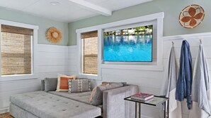 A room with a grey sofa, a small table with a book, a wall-mounted TV, and a large window with a view of a swimming pool. - The Deckhouse | Avila Beach · Steps to Sand (Avila Beach)