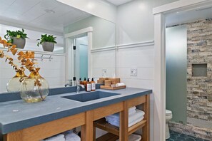 A modern bathroom with a dual sink vanity, a mirror, a wooden shelf with towels, and a shower area with a stone wall.