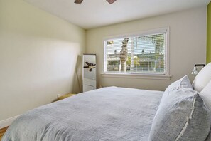 Een slaapkamer met een bed, een raam met zonwering, een plafondventilator en een spiegel.