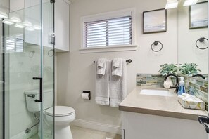 Een badkamer met een douchecabine van glas, een toilet, een wastafel met spiegel en een raam met zonwering.