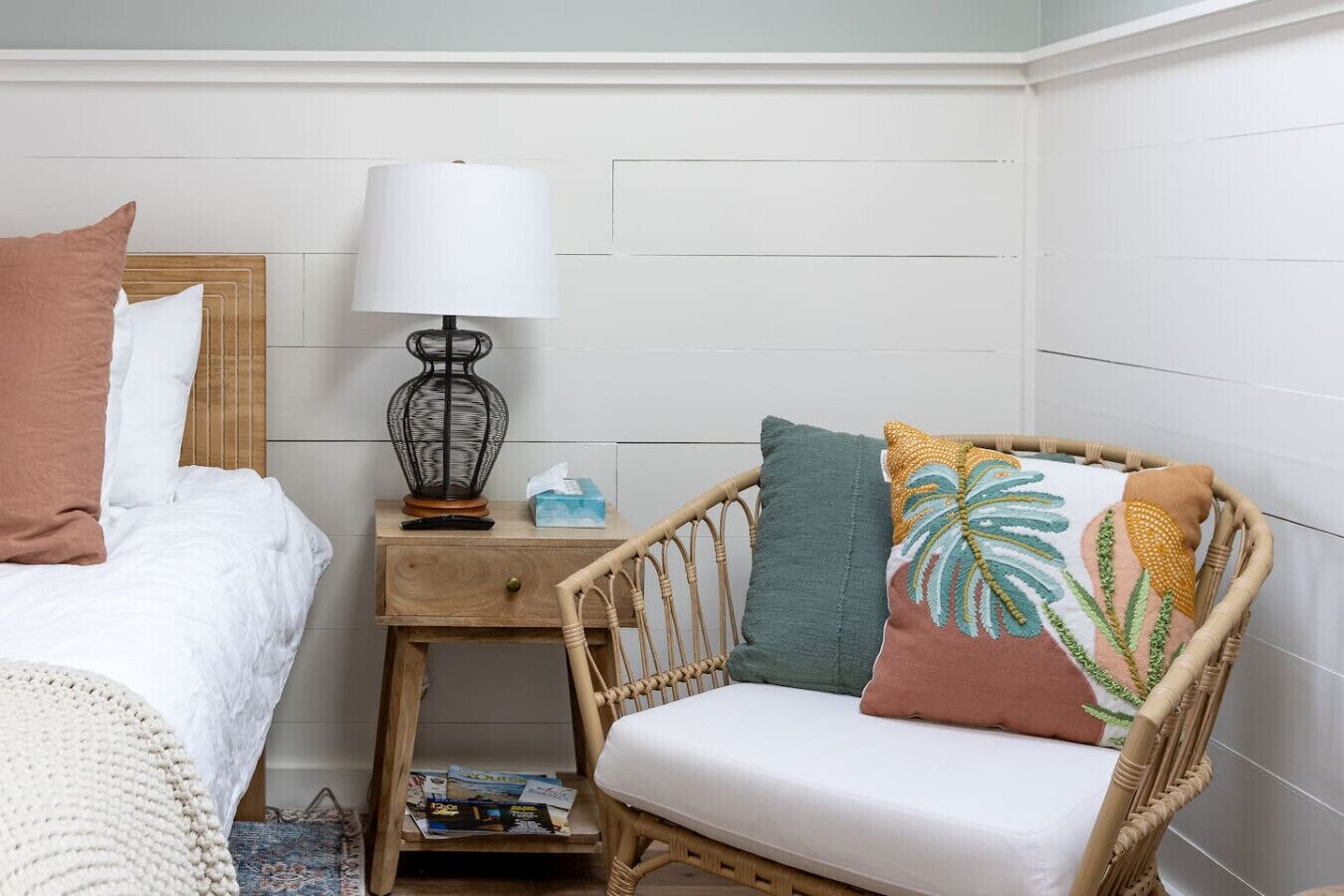 A bedroom with a bed, bedside table, lamp, and wicker chair with cushions.