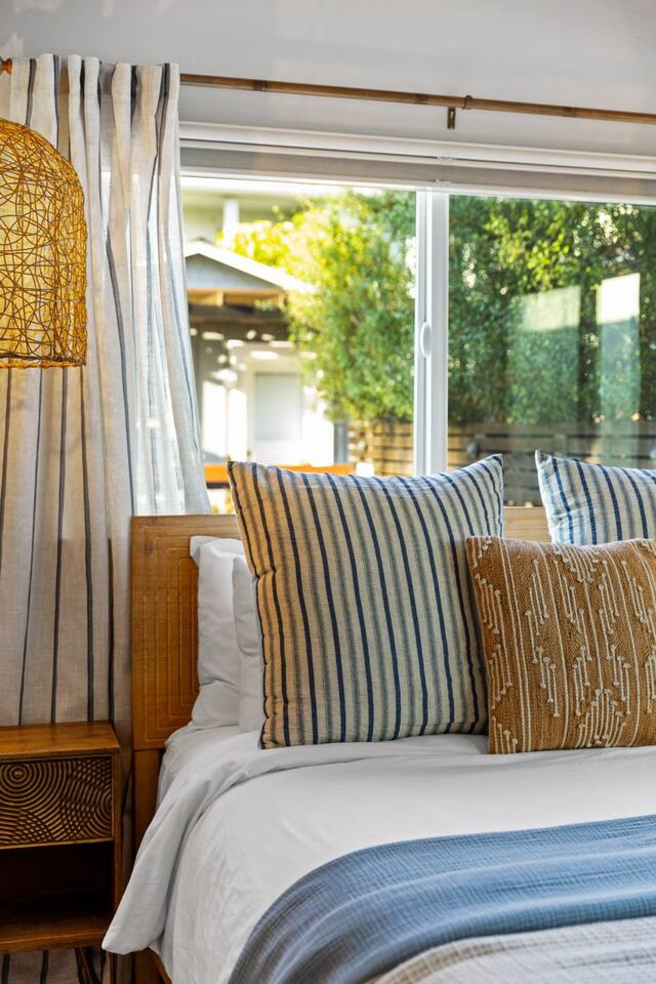 A hotel room with a bed, striped pillows, a wooden nightstand, and a view of greenery outside.