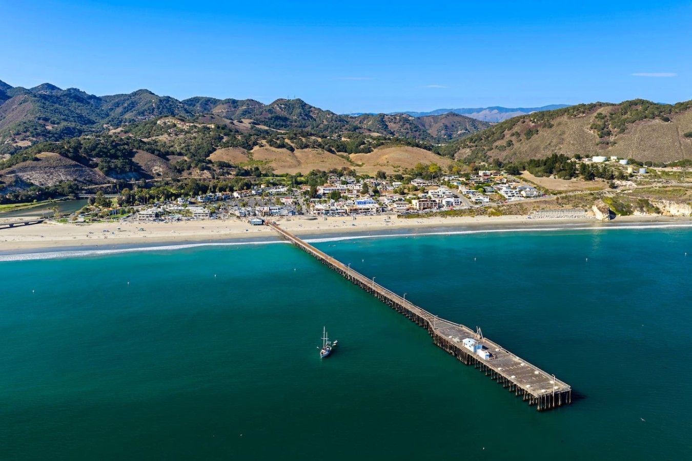 A coastal town with a long pier extending into the sea, surrounded by hills and a beach.