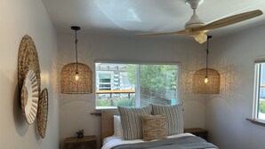 Un dormitorio con una cama, dos lámparas colgantes, un ventilador de techo, una ventana y una mesita de noche con una planta.