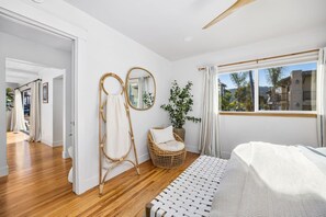 Ein Schlafzimmer mit einem Bett, einem Korbsessel, einem Spiegel und Blick ins Freie.