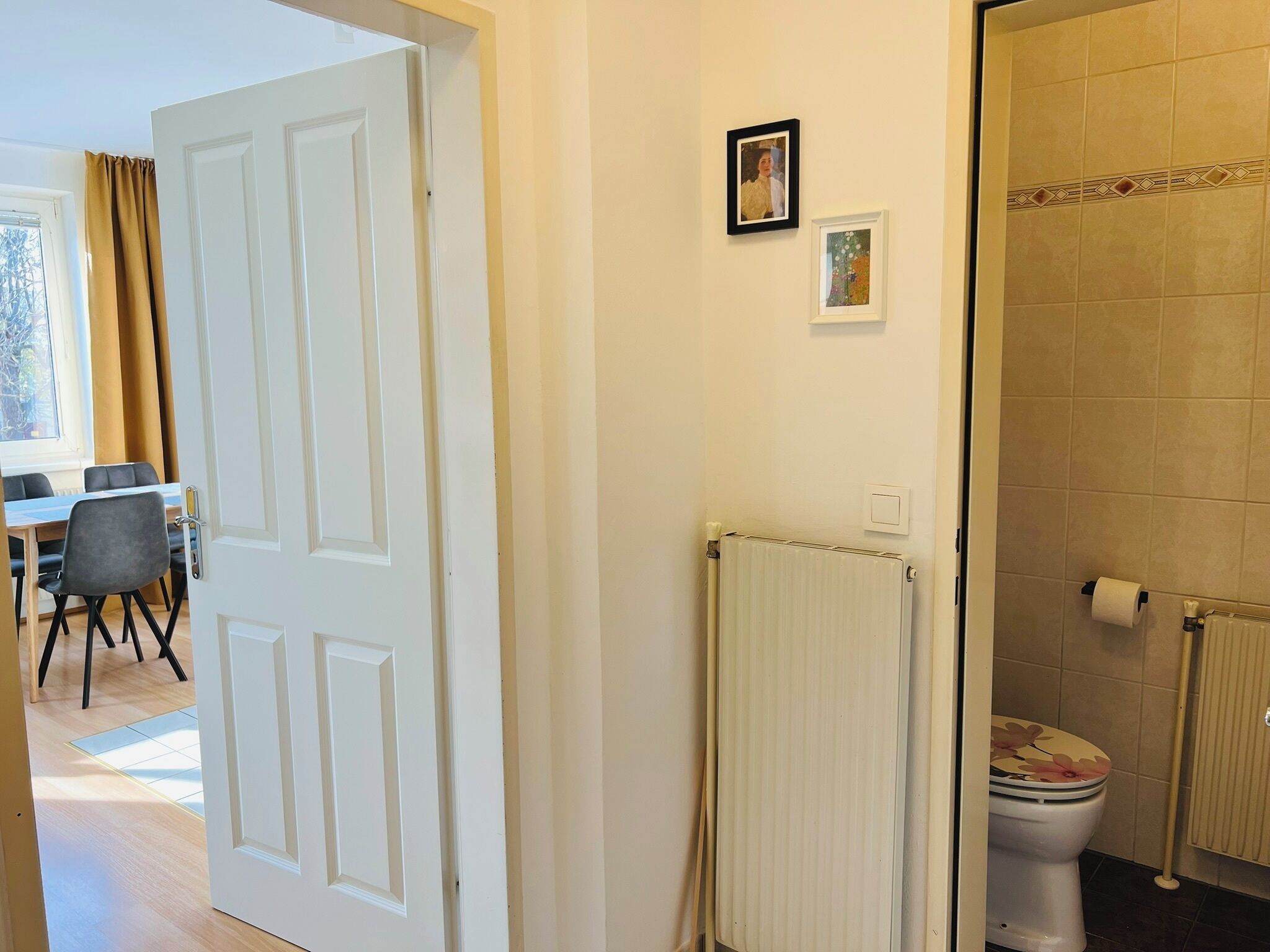 Et værelse med en hvid dør, en radiator, et toilet med blomstret sæde og et lille billedramme på væggen.