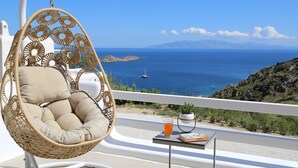 A wicker hanging chair with cushions, a glass of orange juice, and a book on a small table, overlooking a coastal view with a clear sky and a distant sailboat. - Villa Camelia 1 - 1BR - Private Pool & Jacuzzi - Sea View - Psarou (Nammos) (Mykonos)