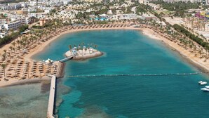 An aerial view of a coastal resort with a central island, a pier, and a beach lined with umbrellas. - HRG Hurghada Hotel Apartments Aqua park-By MAMO (Red Sea Governorate)
