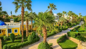 A pathway lined with palm trees and neatly trimmed hedges, leading to a yellow building with blue accents. - HRG Hurghada Hotel Apartments Aqua park-By MAMO (Red Sea Governorate)