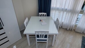 Une petite pièce moderne, avec une table et des chaises blanches, une grande fenêtre ornée de rideaux légers et une vue sur l’extérieur.