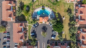 Une vue aérienne d’un quartier résidentiel avec une piscine centrale, entourée de maisons aux toits recouverts de tuiles rouges, et un espace de stationnement où sont garées plusieurs voitures.