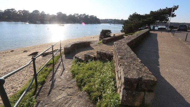 Un sentier de pierres mène à une plage, bordée d’une rambarde et d’un muret, d’où l’on a une vue dégagée sur l’eau.