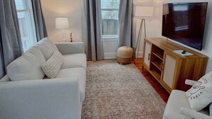A living room with a sofa, ottoman, TV, and a small table with a lamp.
