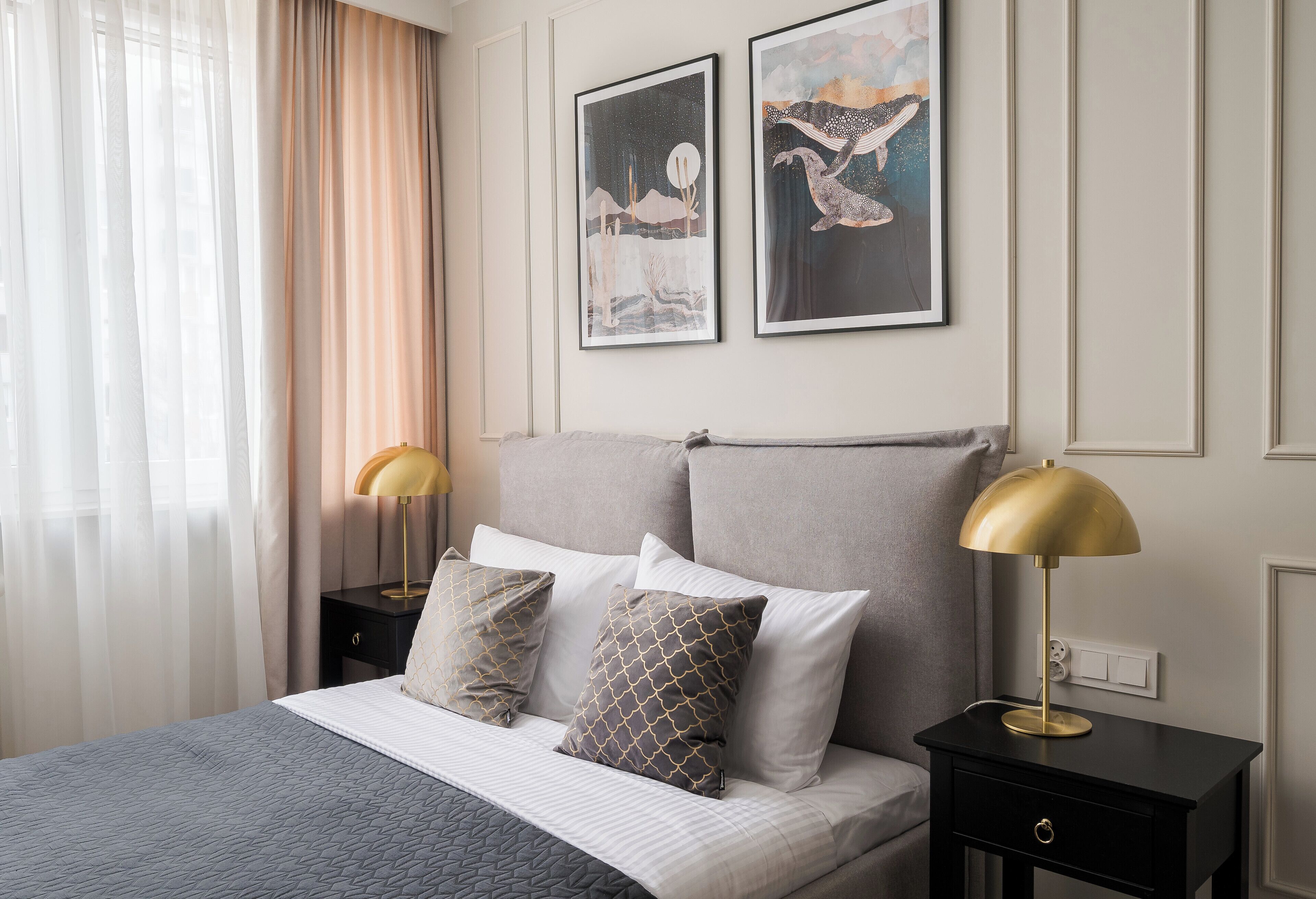 Una cama bien hecha con una colcha gris y blanca, dos almohadas y una lámpara dorada sobre una mesita de noche.