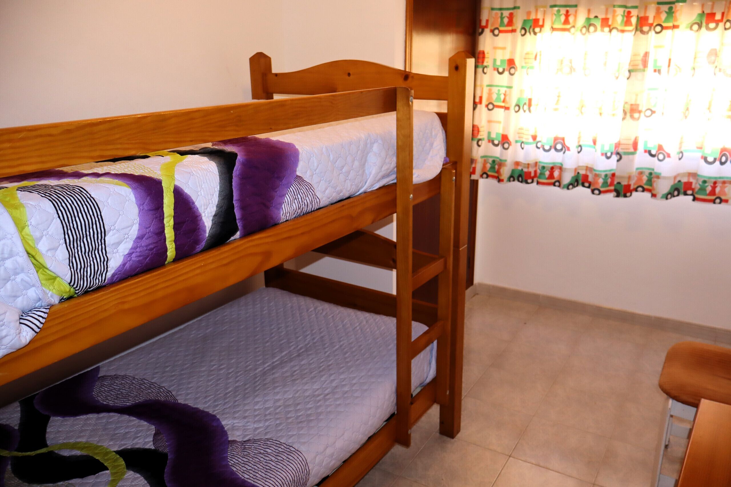 A bunk bed with a purple and white quilt, wooden headboard, and a window with curtains.