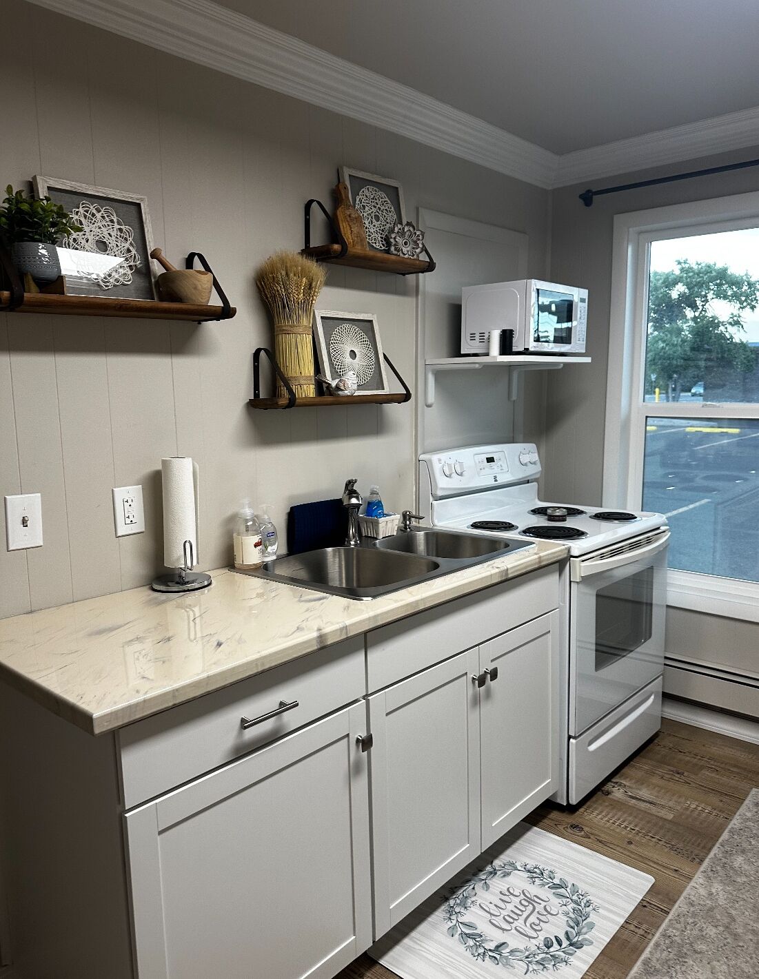 A kitchen with white cabinets, a stove, a microwave, and a sink. There are decorative items on shelves and a window with a view of the outside.