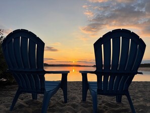 Two Adirondack chairs facing a sunset over a calm lake.