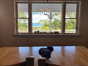 A wooden table with a vase of flowers, a window with a view of greenery and a fence, and a white ceiling light. - “A Gem” 4BR & 2BA—Private Sandy Beach & Bike Path Access (Harwich)