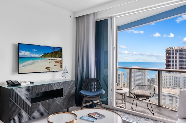 A modern hotel room with a large balcony, a flat-screen TV, a desk, and a comfortable seating area.